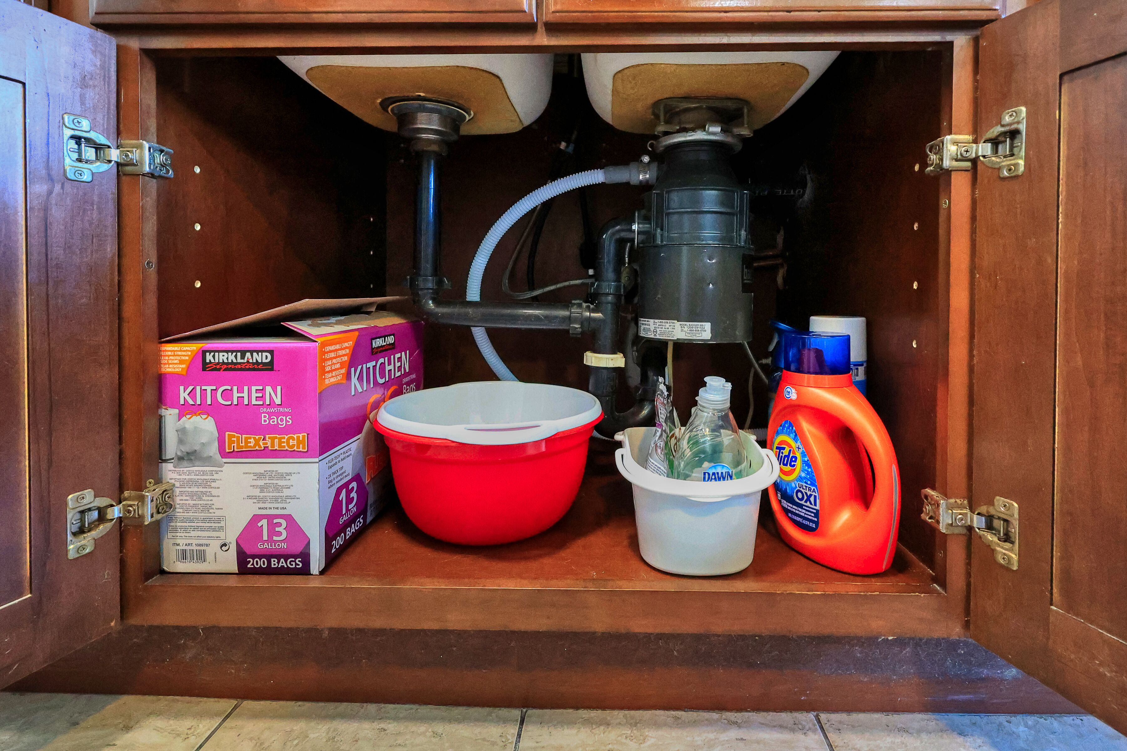A kitchen cabinet with a water filtration system, a red mixing bowl, a white bucket, and cleaning supplies including a box of Kirkland Flex-Tech kitchen bags and a bottle of Dawn dish soap.