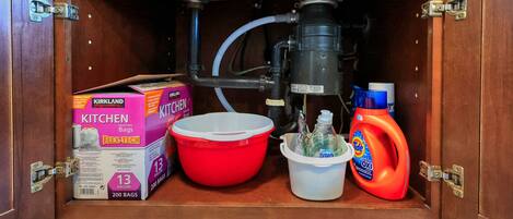 A kitchen cabinet with a water filtration system, a red mixing bowl, a white bucket, and cleaning supplies including a box of Kirkland Flex-Tech kitchen bags and a bottle of Dawn dish soap.