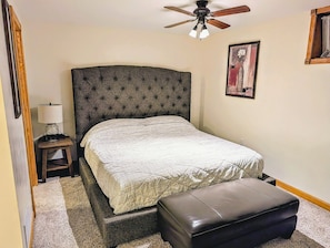 A bedroom with a bed, a nightstand, a lamp, a ceiling fan, and a painting on the wall.