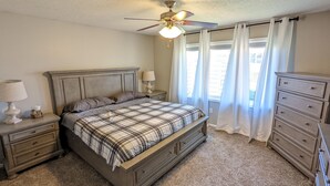Un dormitorio con cama, mesitas de noche, cómoda, ventilador de techo y ventana con cortinas.