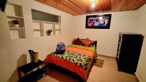 Quarto com teto de madeira, cama com roupa de cama colorida, televisão na parede e uma pequena mesa com uma cesta.