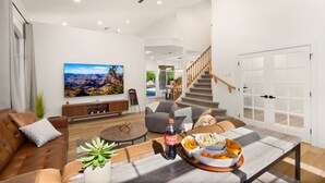 En rummelig stue med en lædersofa, et sofabord med snacks, et stort fladskærms-TV og en trappe, der fører til et andet plan.