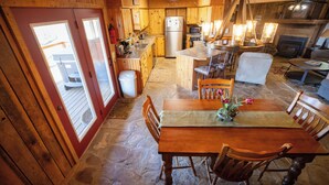 Una cabaña rústica con piso de piedra, interior de madera, cocina con refrigerador y estufa, comedor con mesa y sillas, y chimenea.