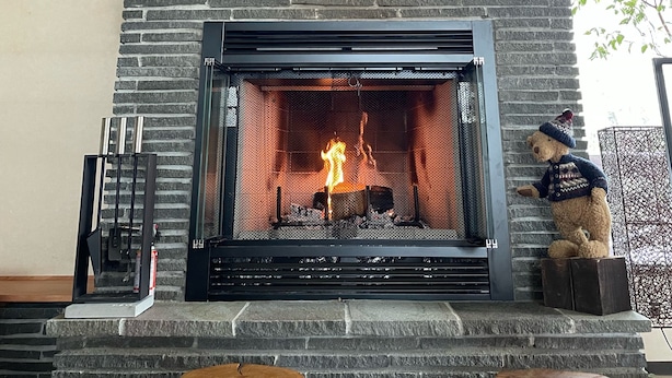 Une cheminée avec une façade en pierre et un foyer en métal, flanquée de deux tabourets en bois et d’un ours en peluche.