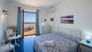 Quarto de hotel com cama, escrivaninha, cadeira, mesa com garrafa e copos, luminária e vista para a praia por uma porta aberta.