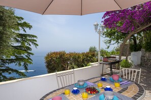 Une table dressée avec des aliments pour le petit-déjeuner, une vue sur la mer et une pergola ornée de fleurs violettes.