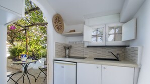 Cozinha compacta com armários brancos, pia e uma pequena geladeira. Há uma mesa de jantar com cadeiras e um enfeite de parede.