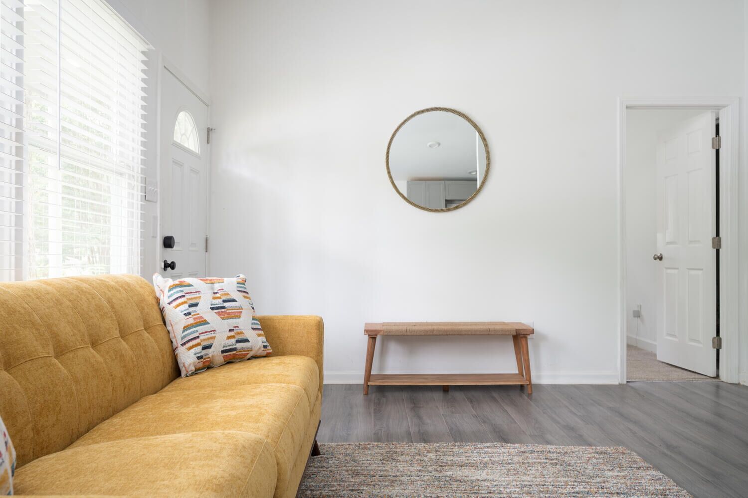 A room with a mustard-colored sofa, a small wooden bench, a round mirror on the wall, and a door to the right.