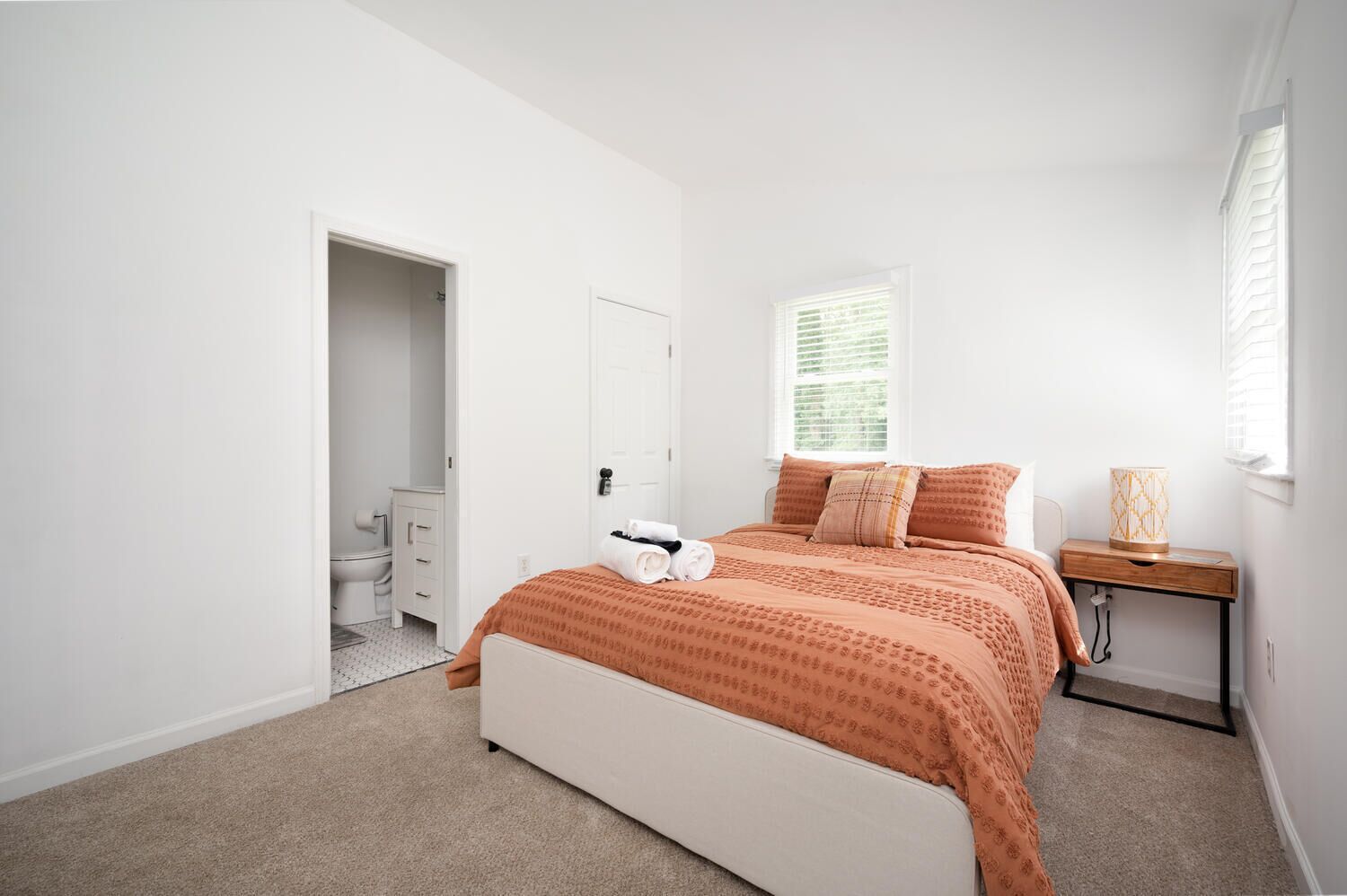 Un dormitorio con cama, mesita de noche, lámpara, ventana y puerta que da a un baño.