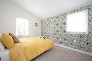 A bedroom with a yellow bedspread, two pillows, a rolled towel, and a patterned wallpaper.