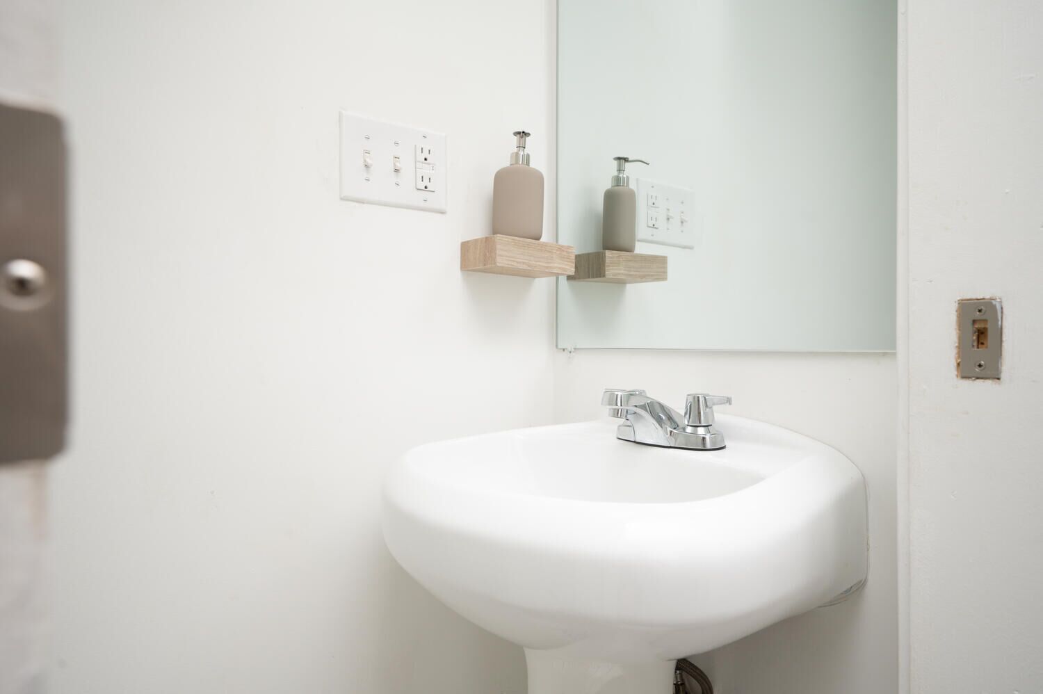 Un baño blanco con lavamanos pedestal, espejo y dispensador de jabón en estante de madera.