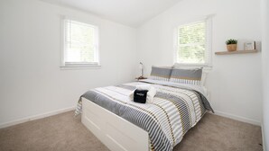 Une chambre à coucher avec un lit, deux fenêtres, une table de chevet, une lampe et une étagère sur laquelle se trouvent une plante et un panneau.