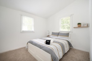 A bedroom with a bed, two windows, a bedside table, a lamp, and a shelf with a plant and a sign.