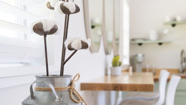 Un vase en métal contenant des tiges de coton et une corde, posé sur une table avec un carnet et des dessous de verre.
