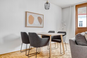 Een moderne eetruimte met een houten tafel en vier grijze stoelen, een gekaderd abstract kunstwerk aan de muur, en een raam met uitzicht op een gebouw.