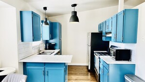 Una cocina con armarios azules, una encimera blanca, un refrigerador negro, un microondas y una lámpara colgante.