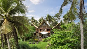 A two-story house with a thatched roof, surrounded by lush greenery and palm trees. - The Cove Bali by Nakula (Selemadeg)