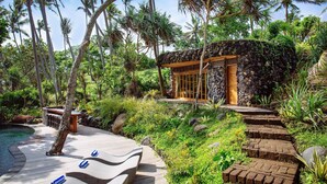 A tropical resort with a stone-walled lounge area, a wooden deck, and a stone staircase leading to a wooden structure. - The Cove Bali by Nakula (Selemadeg)