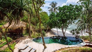 A tropical pool area with lounge chairs, surrounded by lush greenery and a beach view. - The Cove Bali by Nakula (Selemadeg)