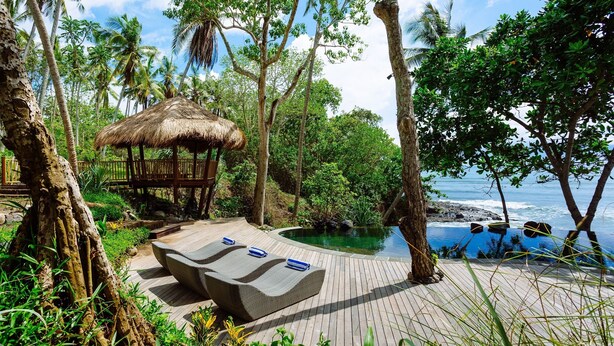 A wooden deck with lounge chairs, a thatched-roof structure, and a pool surrounded by tropical vegetation and palm trees. - The Cove Bali by Nakula (Selemadeg)