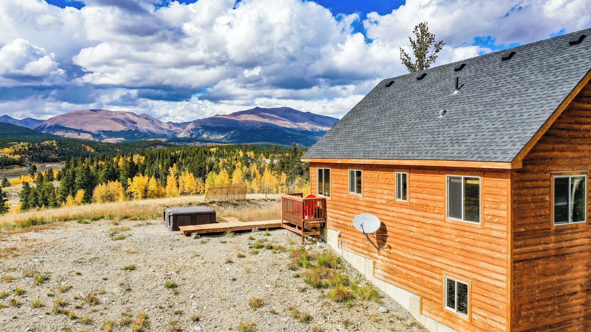 A wooden cabin with a satellite dish, a deck, and a view of mountains and autumn trees.
