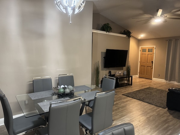 A modern dining area with a glass table and grey chairs, a flat-screen TV mounted on the wall, and a wooden door in the background.