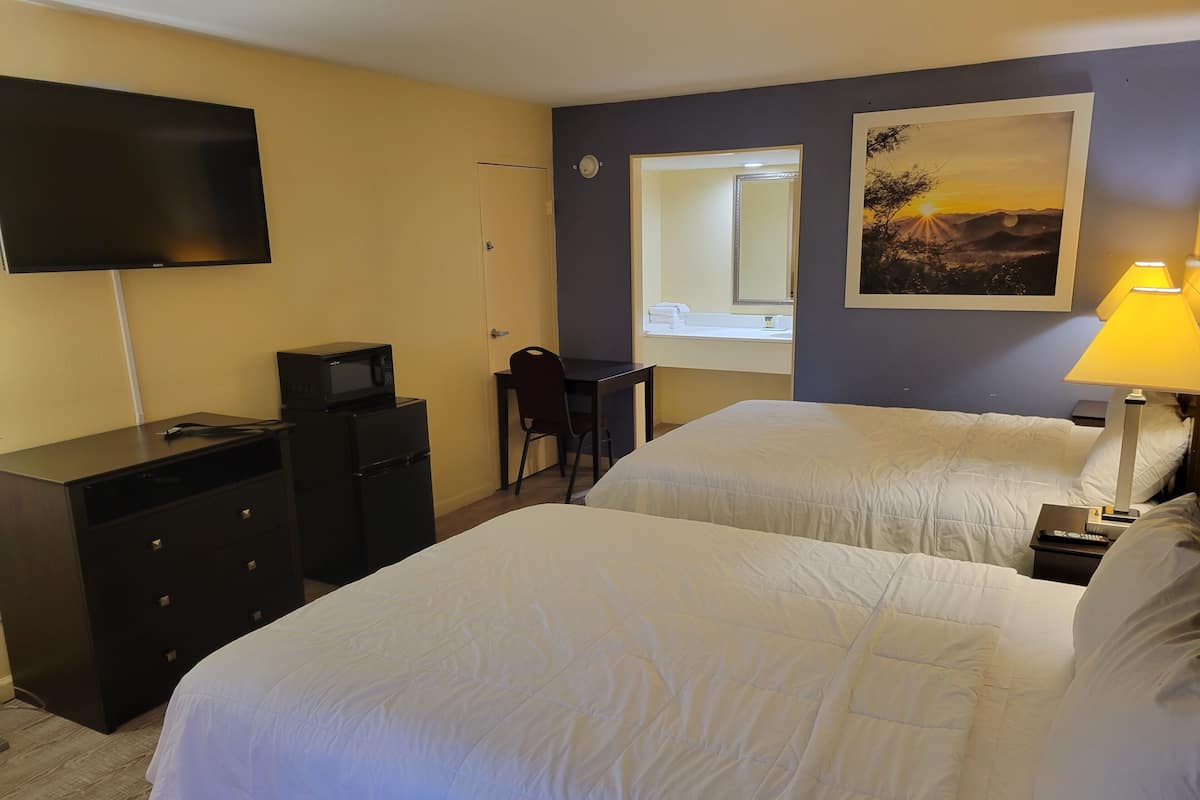 A hotel room with two beds, a desk, a TV, a microwave, a chair, a lamp, and a painting on the wall.