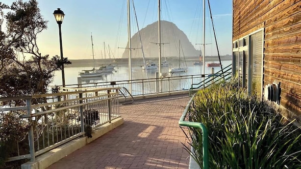 A waterfront walkway with a wooden building on the right, leading to a marina with boats and a mountain in the background.