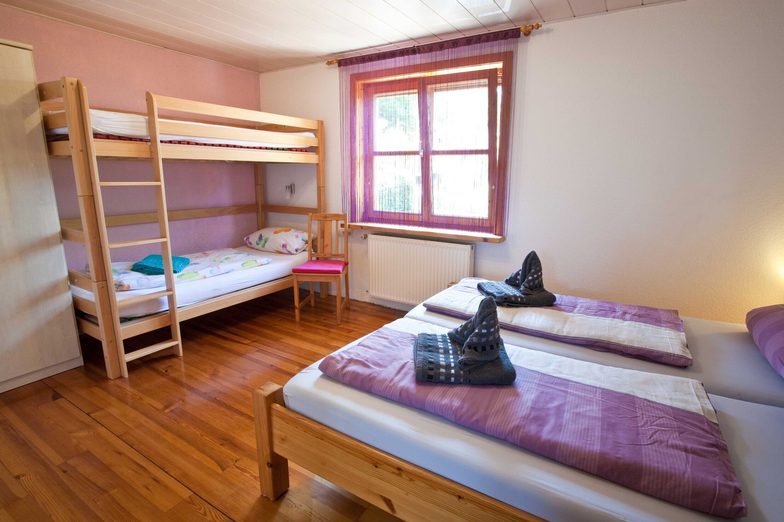 A dormitory room with bunk beds, a wooden ladder, a radiator, and a window with curtains.