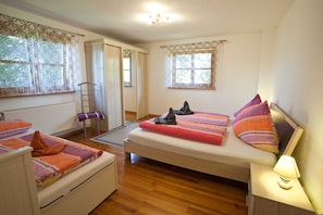 A bedroom with a bed, a bedside table with a lamp, a window with curtains, a radiator, and a door leading to another room.
