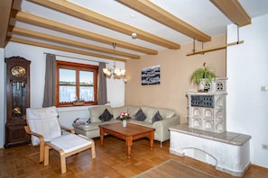 A living room with a wooden ceiling, a sofa, a coffee table, a chair, a fireplace, a clock, and a window. - Large Apartment in Allgäu with Garden & Wi-Fi (Weiler im Allgäu)