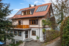 A two-story house with a sloped roof, large windows, and a wooden balcony. - Large Apartment in Allgäu with Garden & Wi-Fi (Weiler im Allgäu)