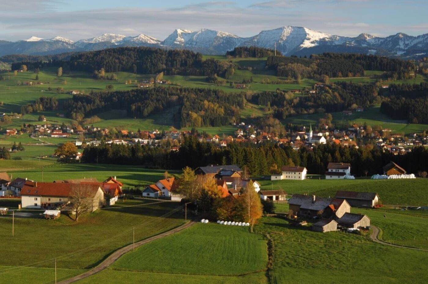 Een landelijk landschap met een dorp, groene velden en besneeuwde bergen op de achtergrond.