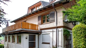 A two-story house with a wooden balcony, a garage door, and a garden area.