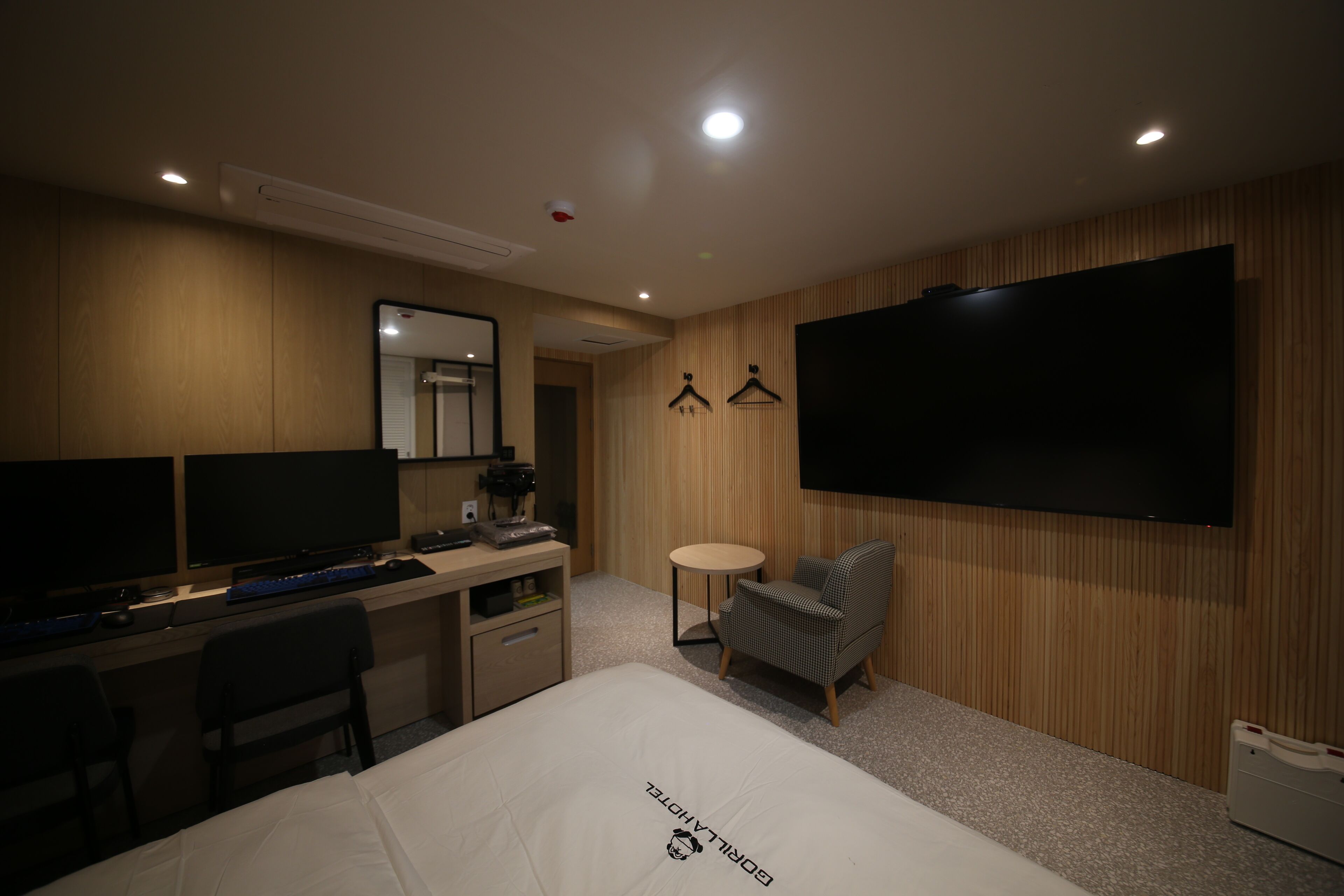 Habitación de hotel con una cama, escritorio, dos monitores, una silla, una mesita y un televisor.