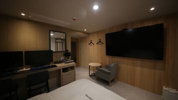 Habitación de hotel con una cama, escritorio, dos monitores, una silla, una mesita y un televisor.