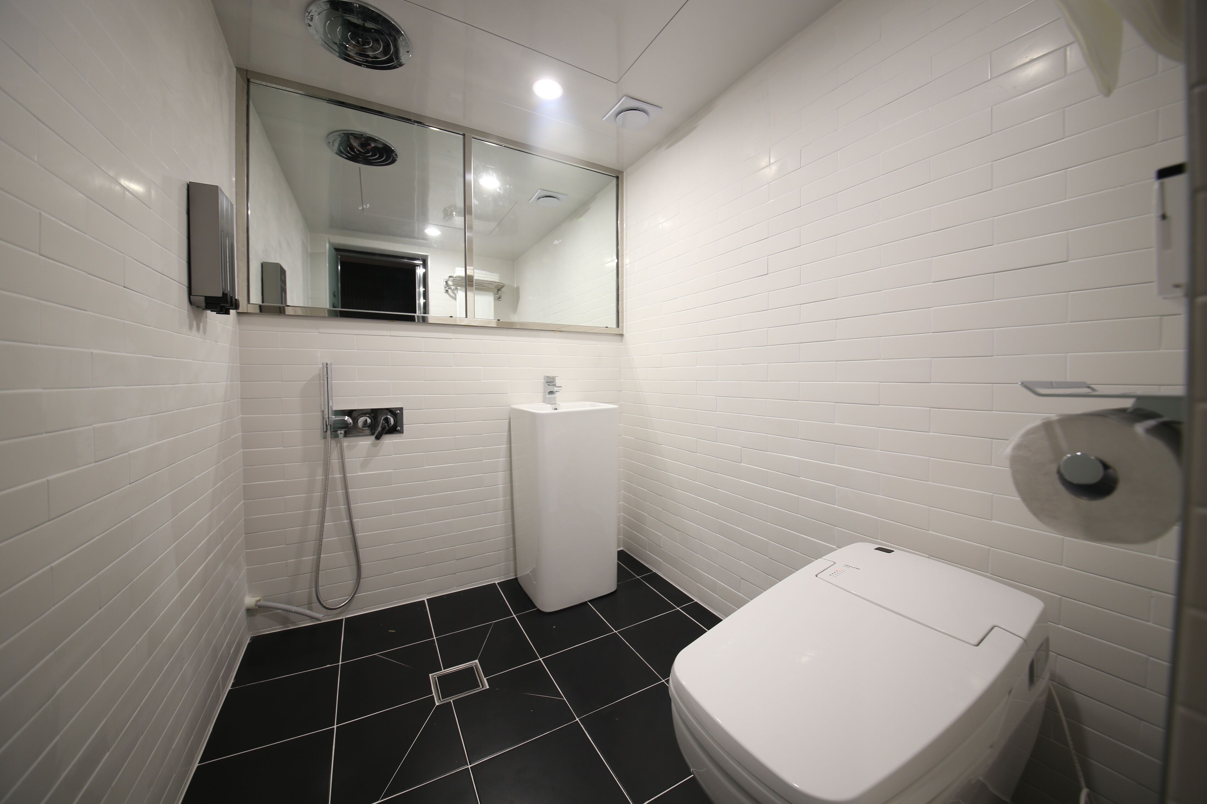 A small, modern bathroom with white tiled walls and a black tiled floor. It includes a toilet, a sink, and a shower area.