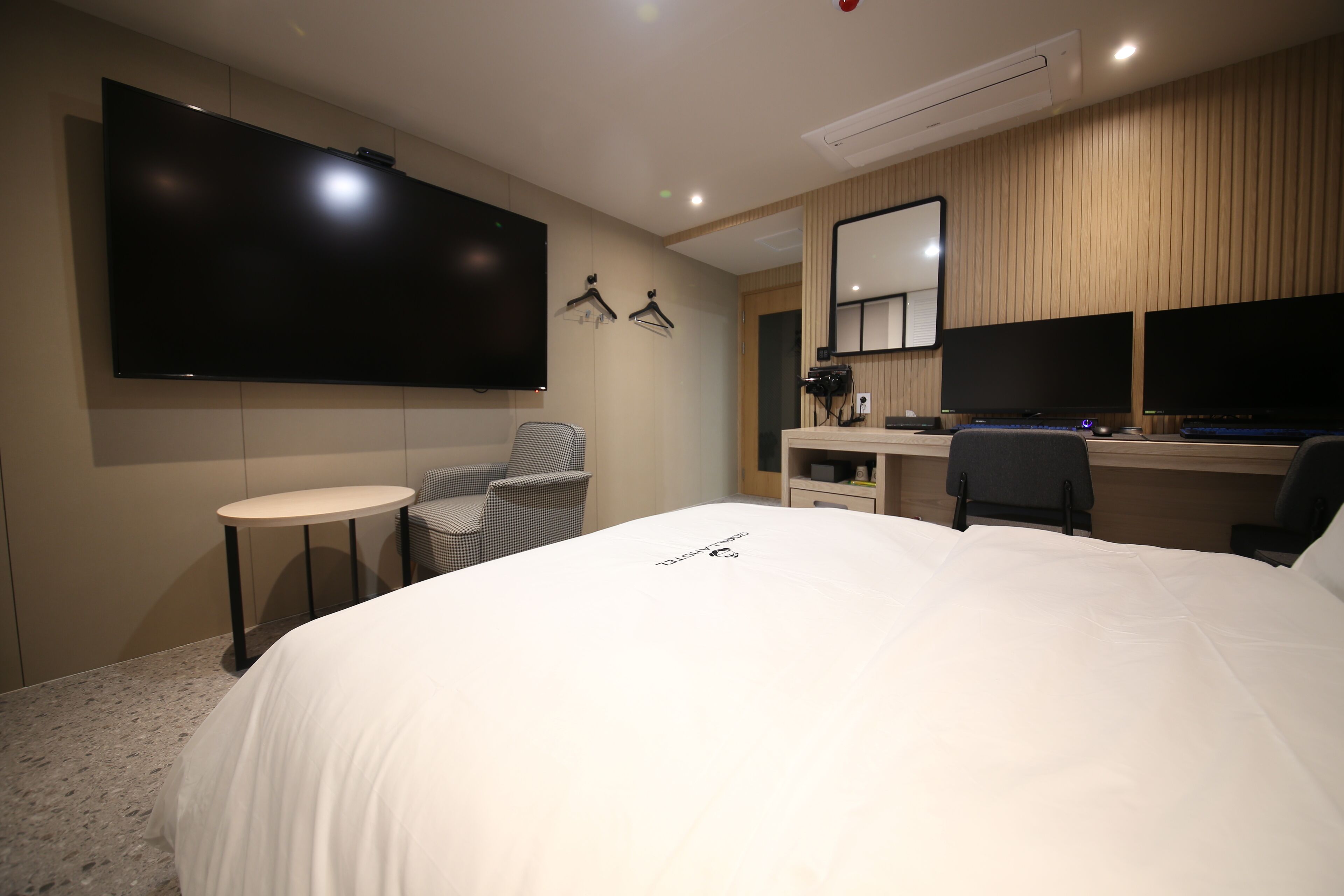 A hotel room with a large bed, a desk with two monitors, a TV mounted on the wall, and a small round table.