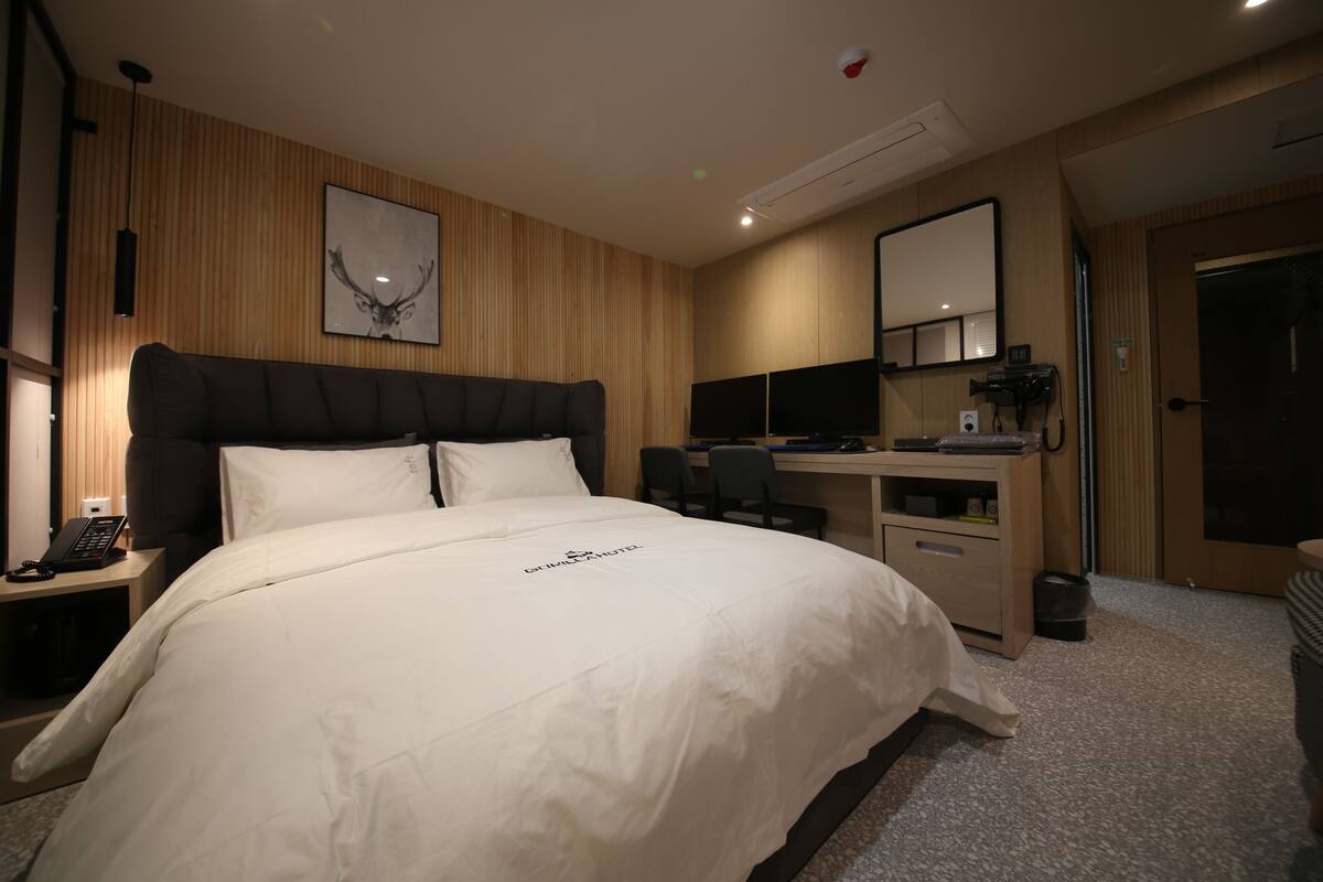 Une chambre d’hôtel avec un grand lit, un bureau, deux écrans d’ordinateur, un téléphone et un miroir.