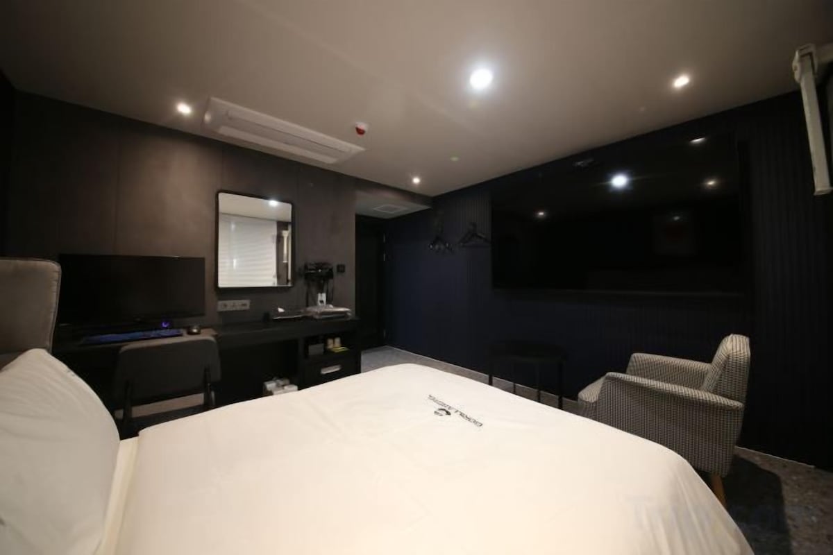 Ein Hotelzimmer mit einem großen Bett, einem Schreibtisch, einem Sessel, einem Fernseher und einer dunkel gestrichenen Wand.