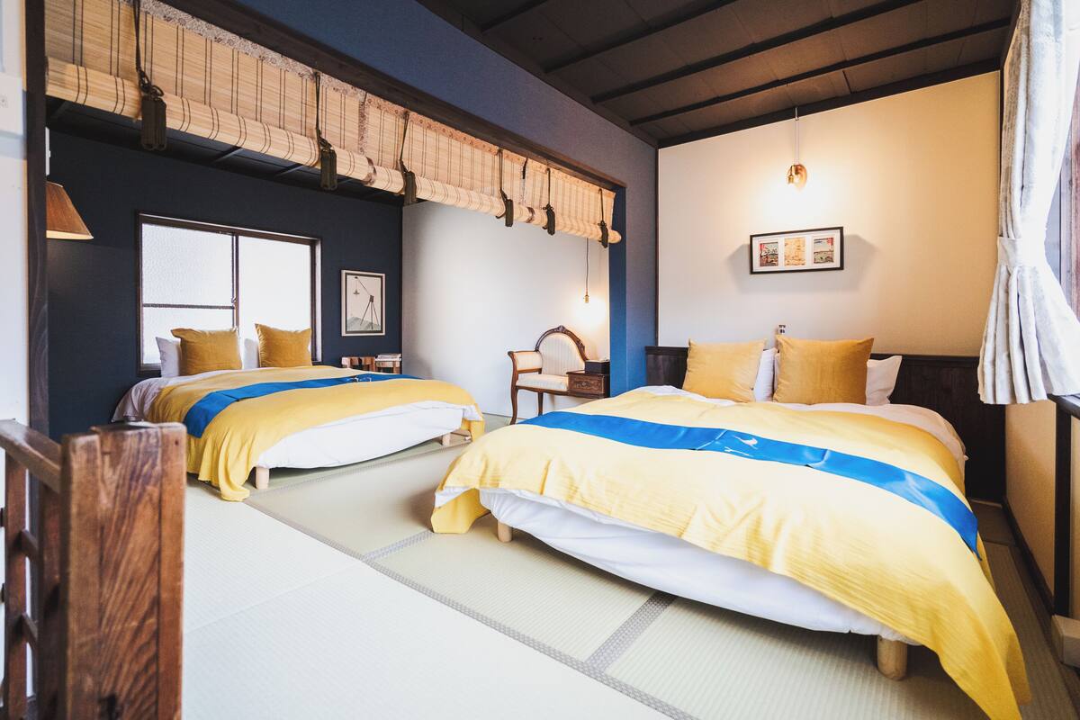 A traditional Japanese-style room with tatami flooring, two beds with yellow and blue bedding, a wooden ceiling, and a hanging shoji screen.
