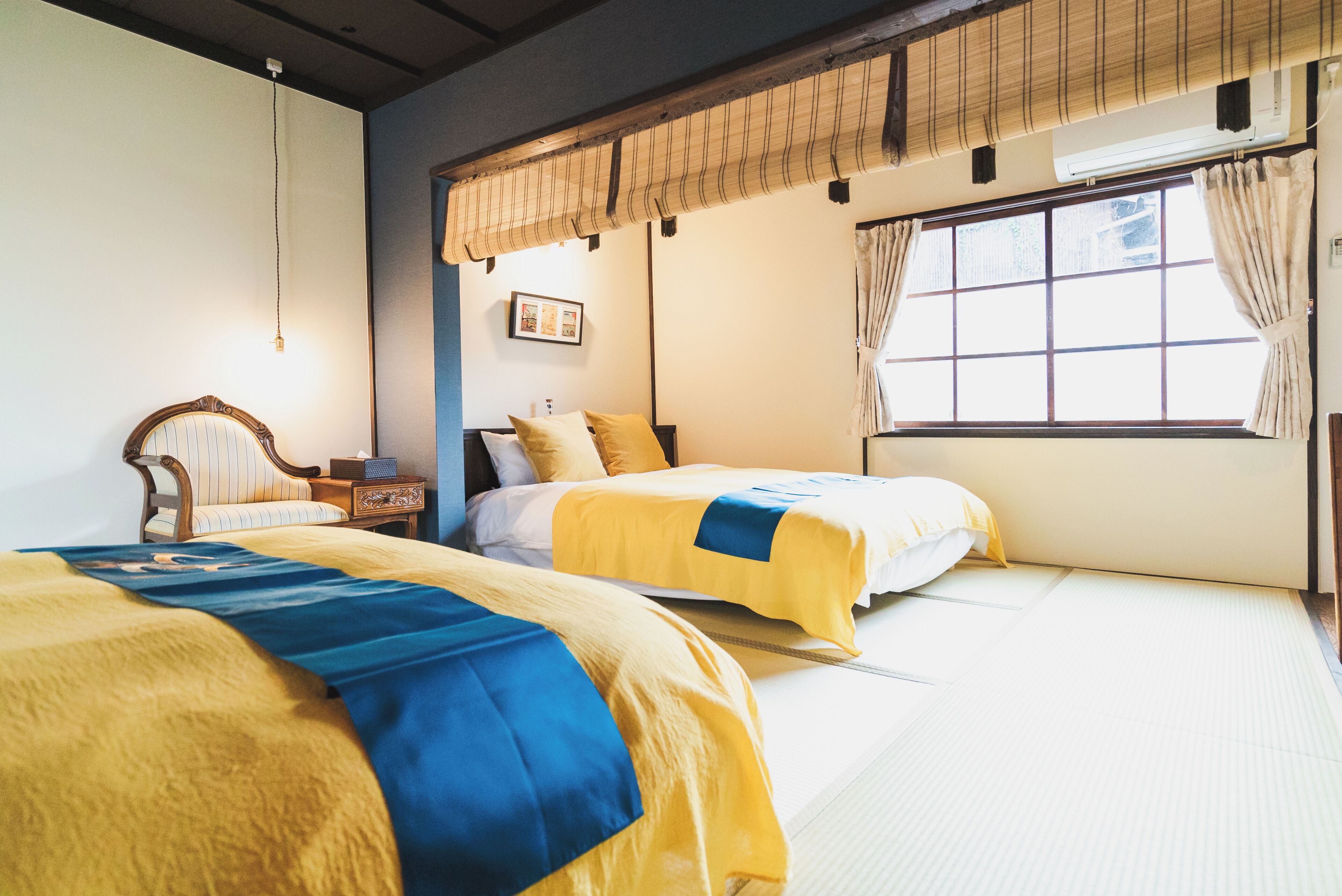 Una habitación tradicional japonesa con suelo de tatami, dos camas con ropa de cama amarilla y azul, una silla de madera y una ventana con cortinas.
