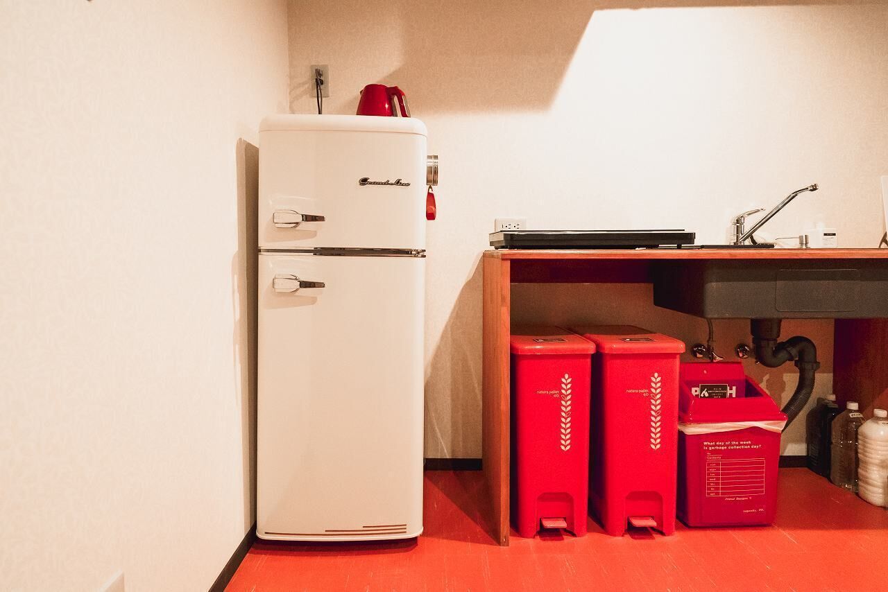 Une petite cuisine équipée avec un réfrigérateur blanc, une machine à café rouge et deux poubelles rouges.