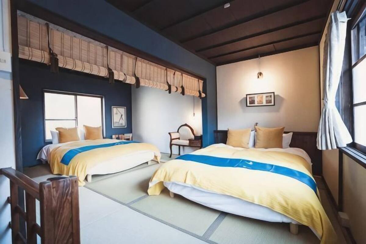 A room with two beds, a chair, a window, and a wall-mounted light fixture.
