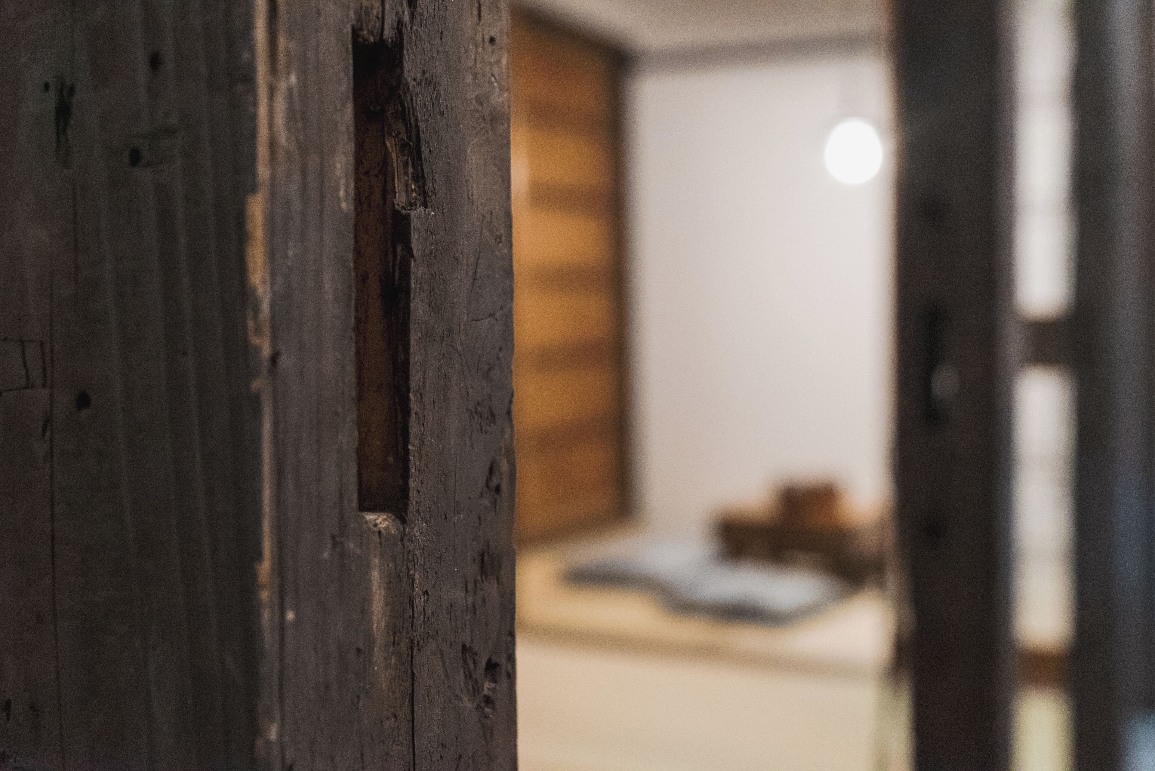 A wooden door with a visible gap, leading to a room with a bed and a wooden wall.