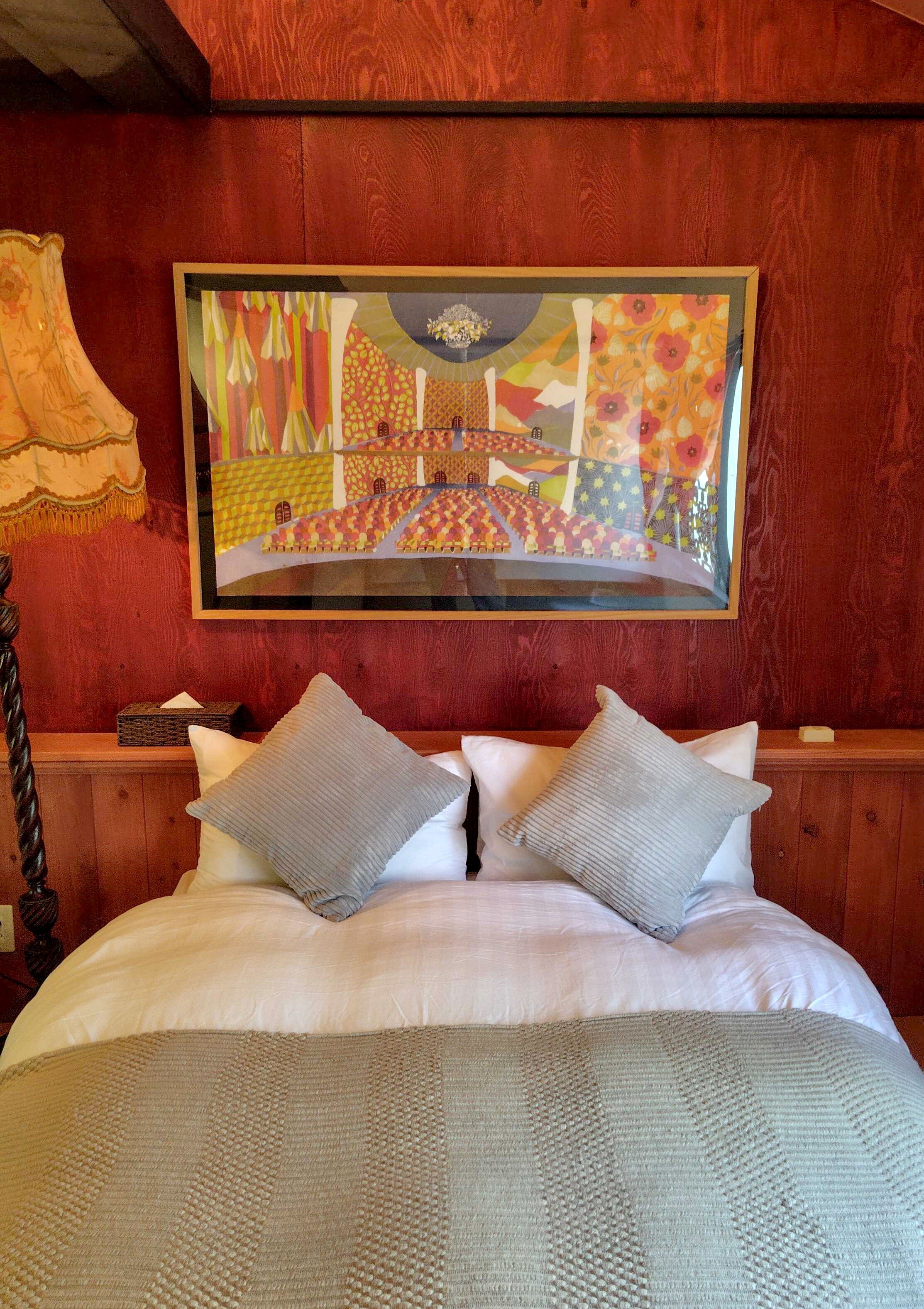 Un lit avec une couverture à motifs et des oreillers, une tête de lit en bois et une œuvre d’art encadrée au-dessus du lit.