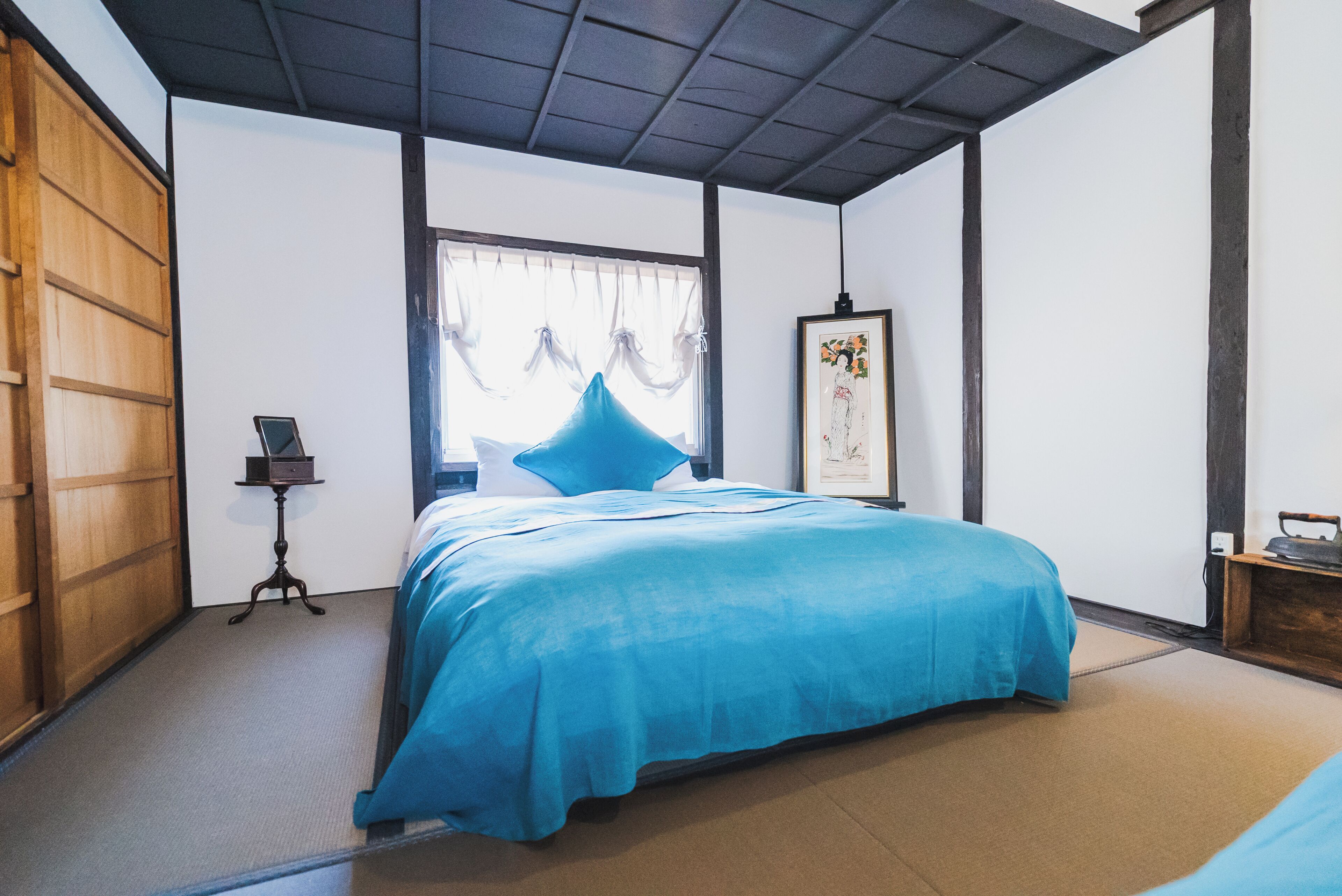 Une pièce de style japonais traditionnel, avec un sol recouvert de tatamis, un lit avec une couvre-lit bleue, un paravent shoji et une œuvre d’art encadrée accrochée au mur.