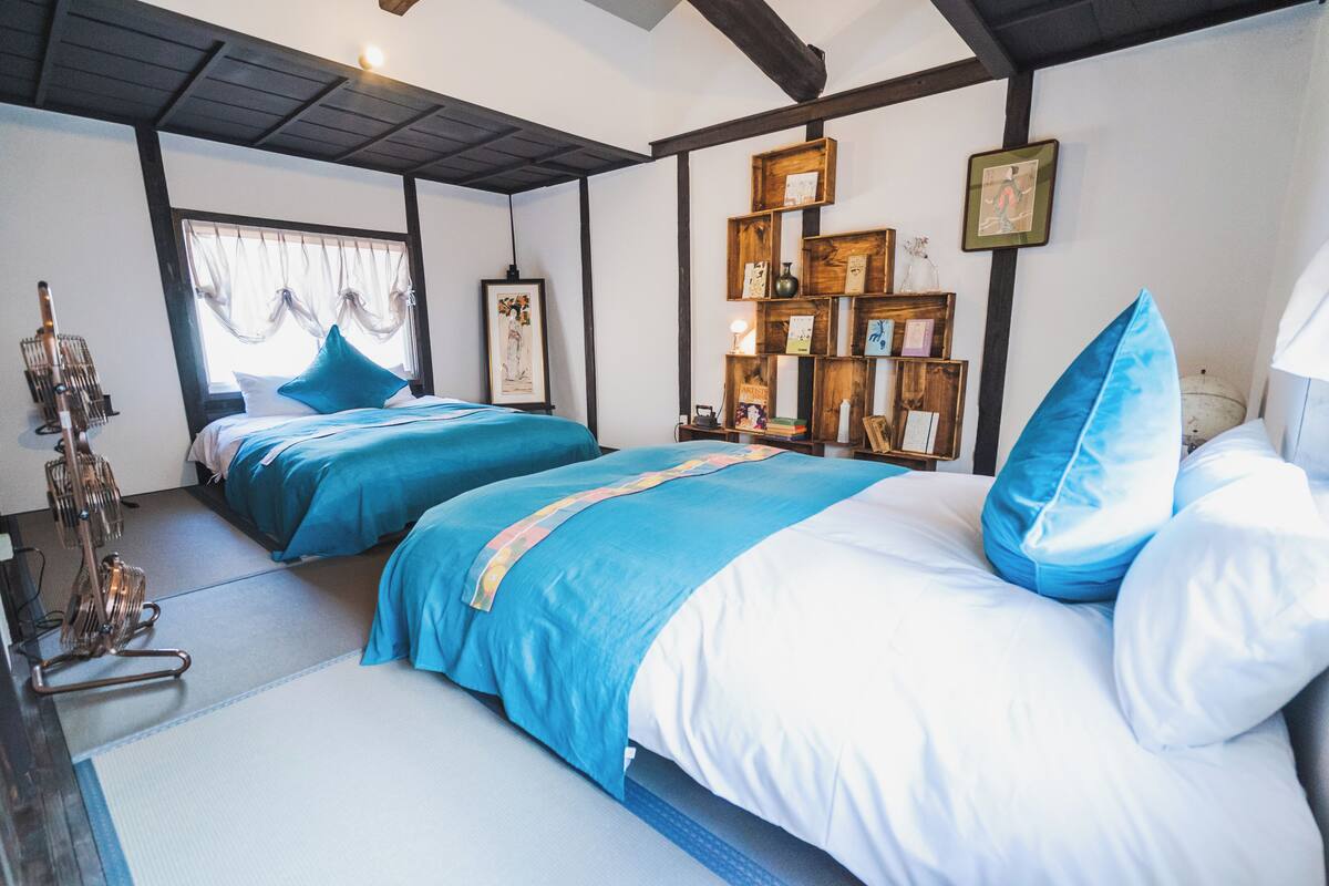 A hotel room with two beds, a wooden shelf with framed pictures, a small table, and a window with curtains.