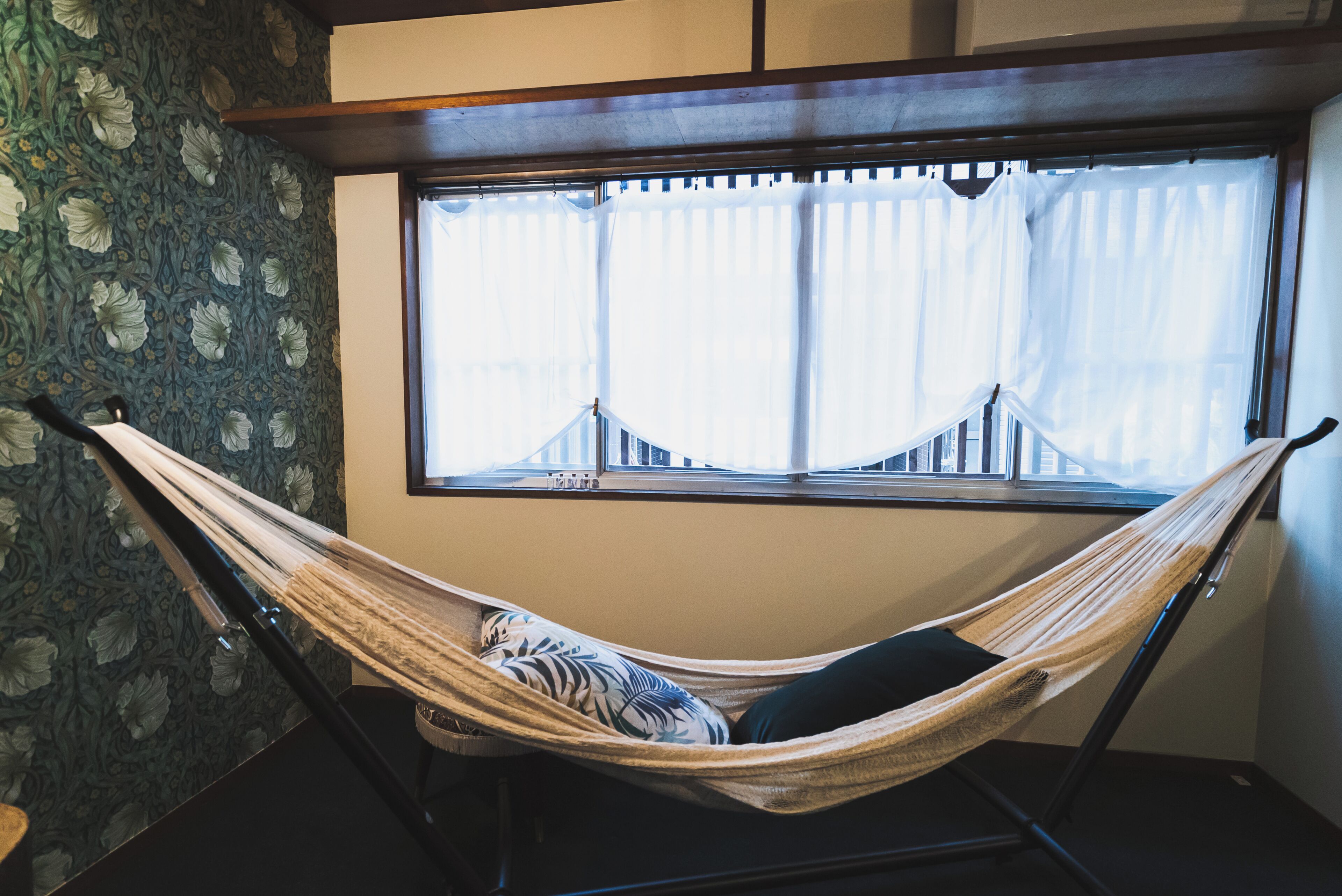 A hammock in a room with floral wallpaper and a window with sheer curtains.