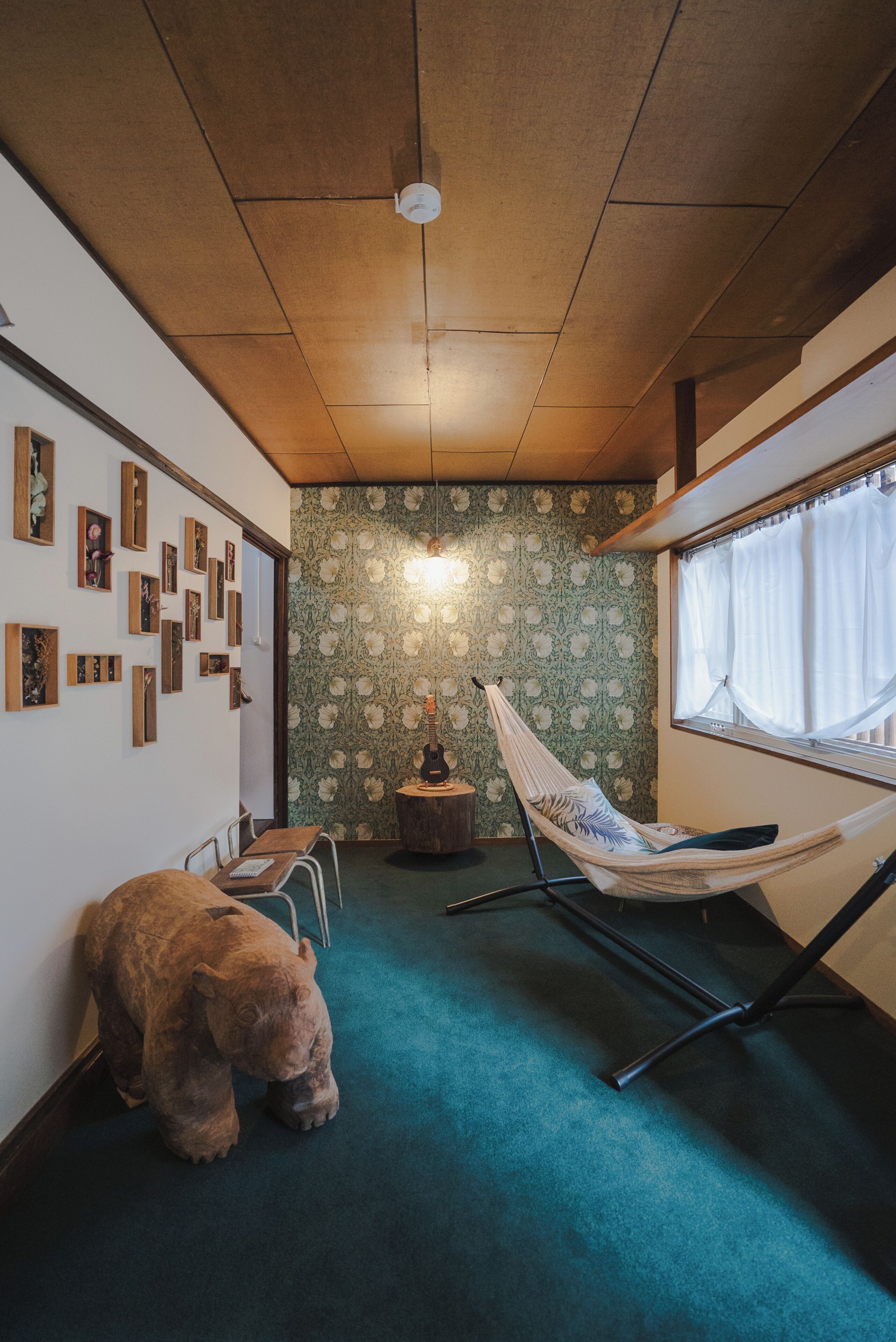 A room with a wooden ceiling, a hammock chair, a small table, a teddy bear, and a wall with framed pictures.